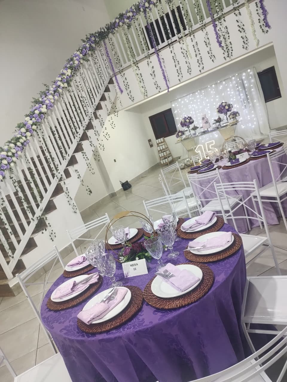 Decoração de festa de 15 anos com mesa redonda lilás e escada enfeitada de flores