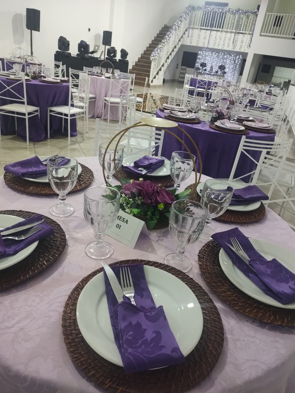 Salão com mesas redondas em toalhas lilás e roxas e escada decorada ao fundo