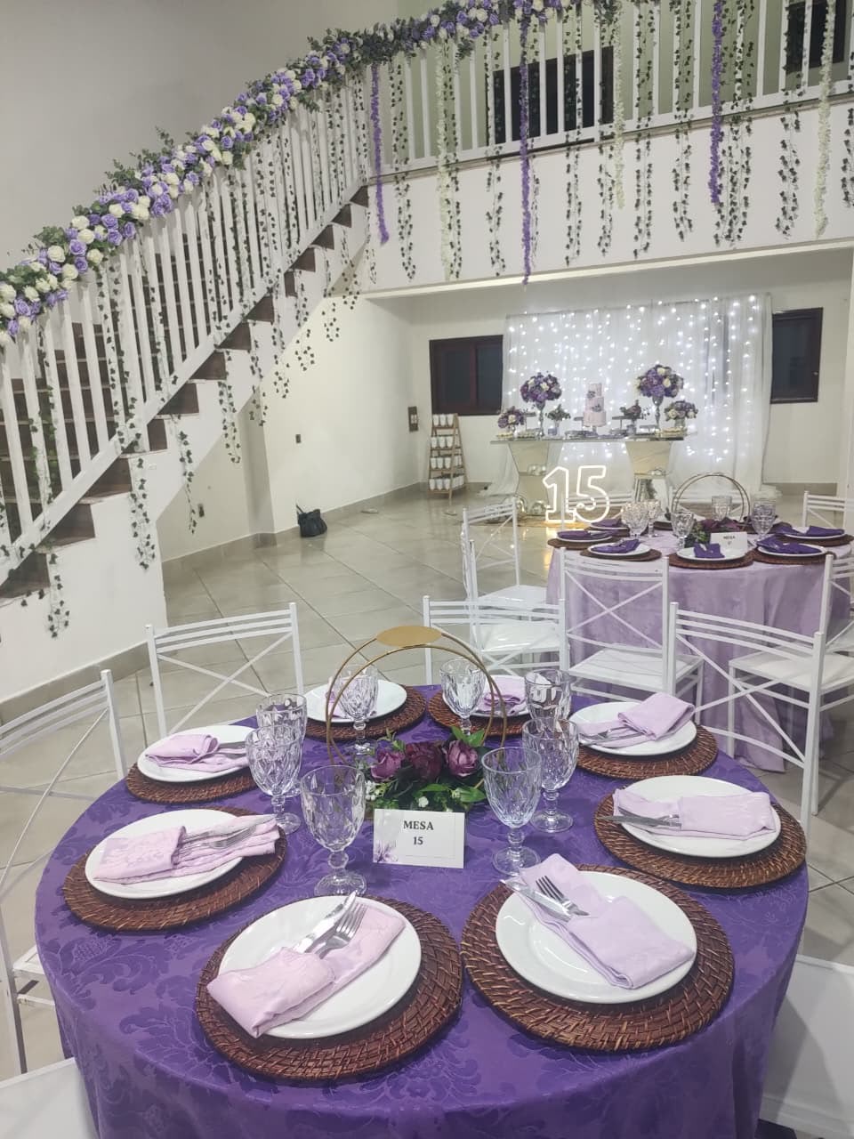 Salão montado com mesas lilás, escada decorada com flores e mesa de doces ao fundo
