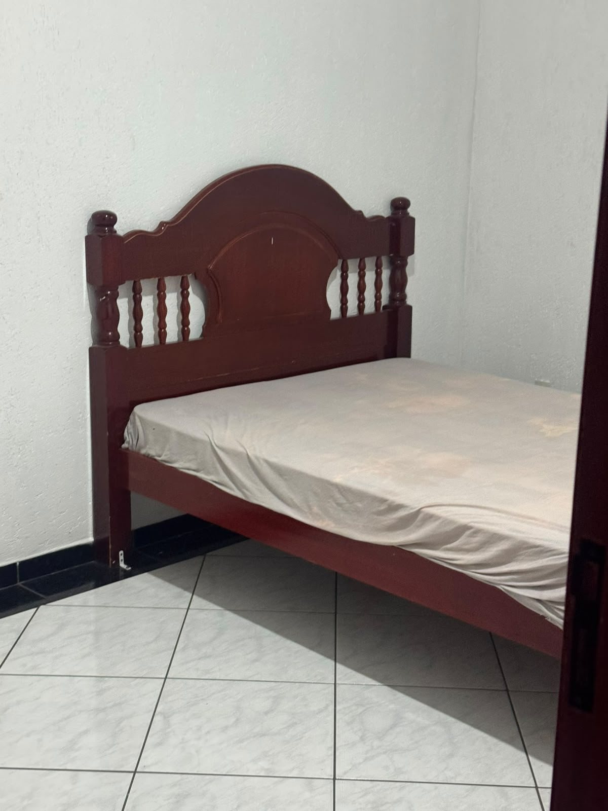 cama de madeira escura com cabeceira entalhada e colchão com lençol bege num quarto com parede branca e piso de cerâmica clara  — texto: